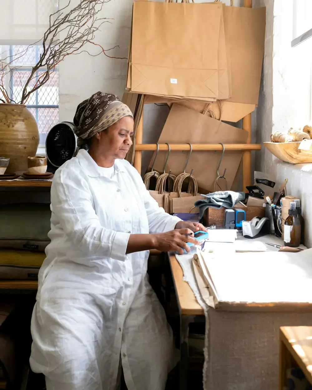 A woman in white working at a desk in a bright craft studio, paper bags on a rack behind her