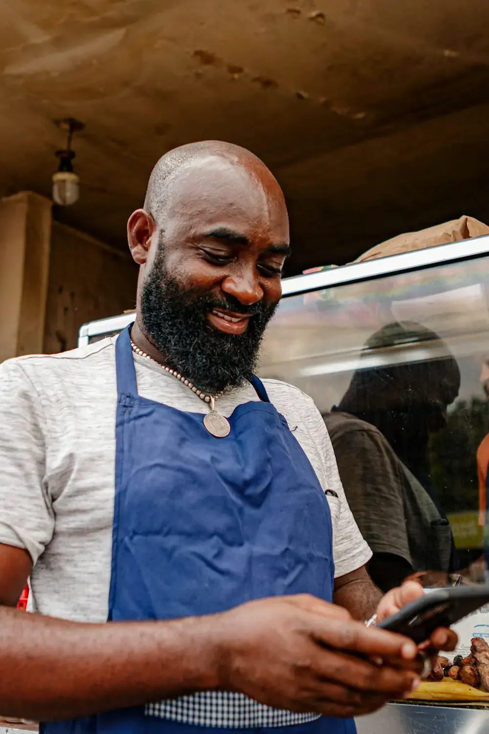 A smiling man in a blue apron looking at his phone at a food counter