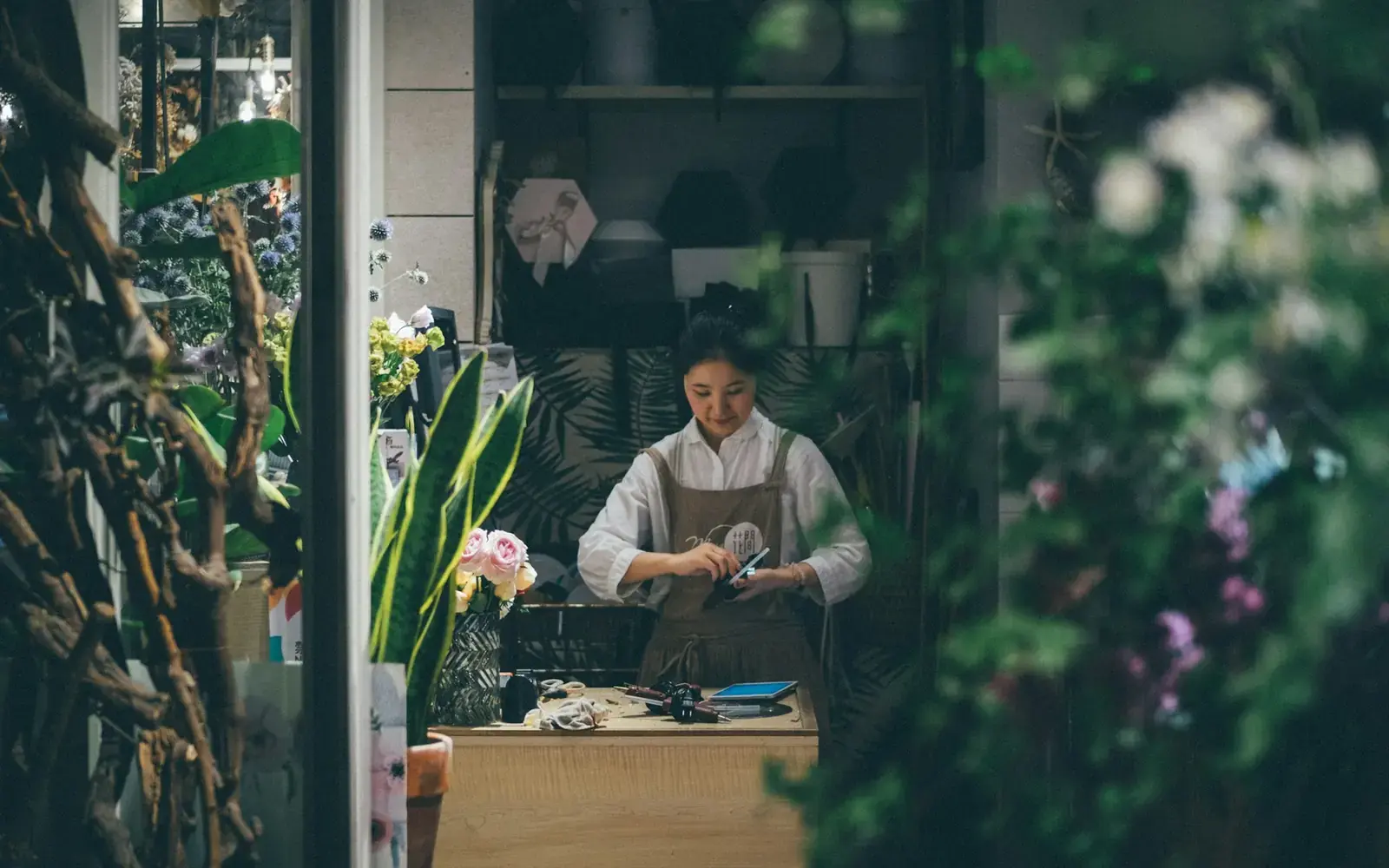 A florist wrapping an order at her workbench, seen through the shop window